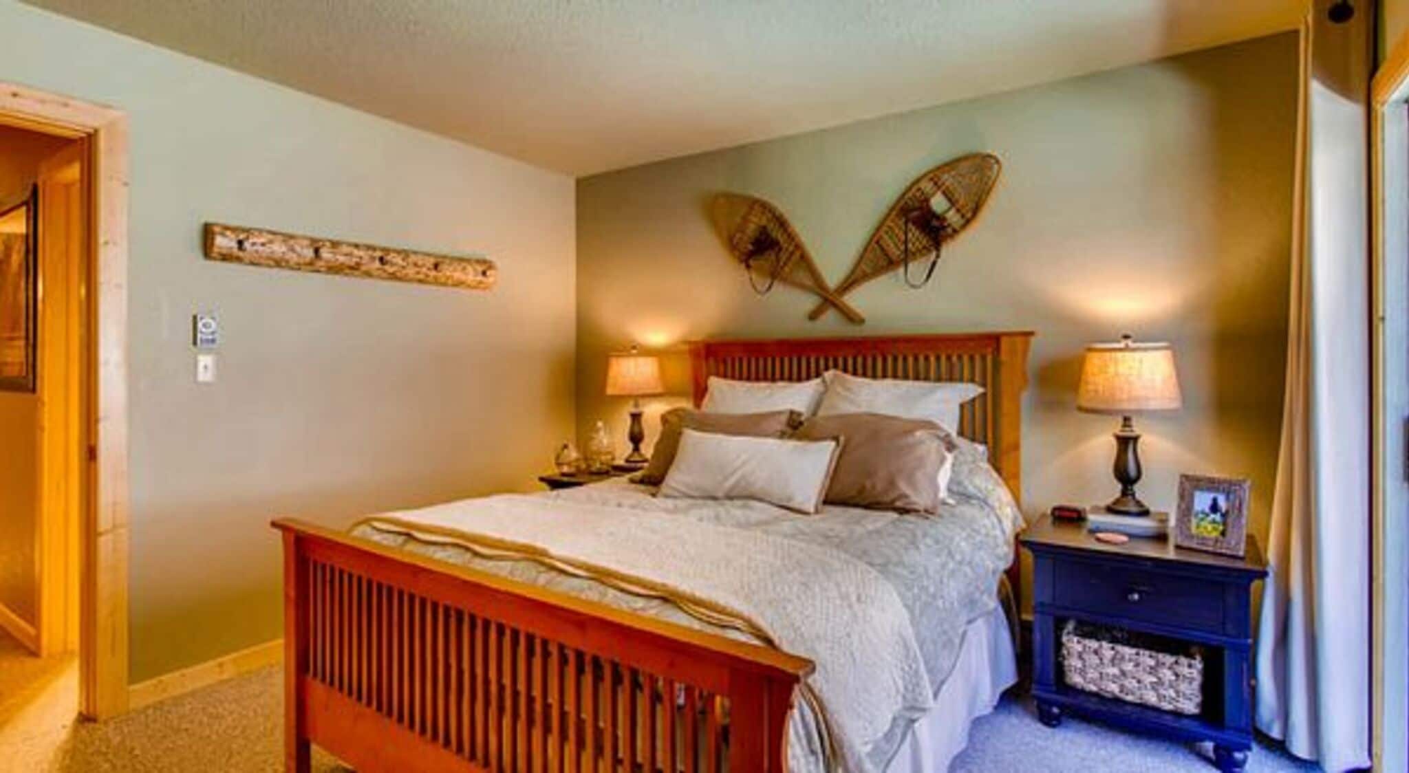 Quarto com cama de madeira, duas luminárias de cabeceira, mesa de apoio e um objeto decorativo na parede.