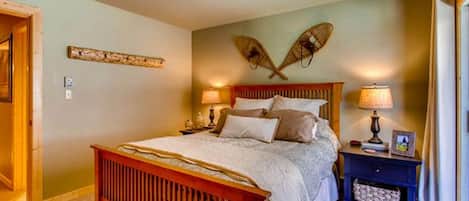 Quarto com cama de madeira, duas luminárias de cabeceira, mesa de apoio e um objeto decorativo na parede.