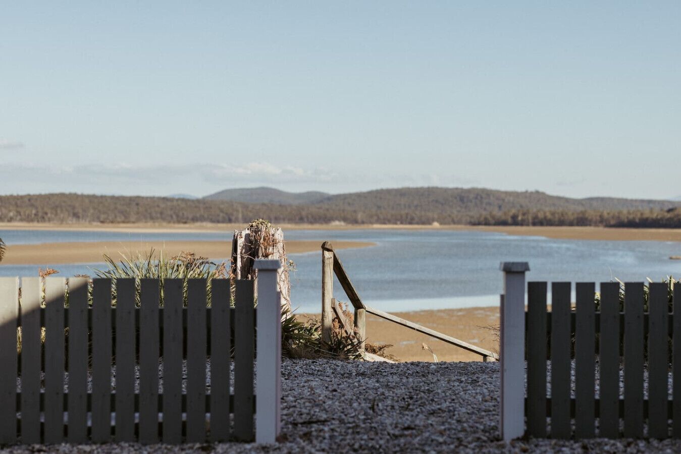 A wooden gate leading to a beach with a clear view of the water and distant hills.