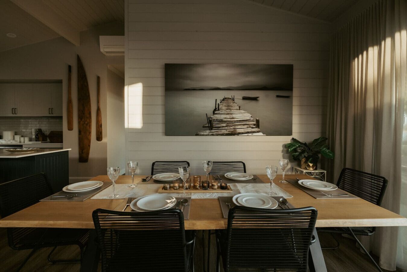 A dining area with a wooden table set for six, surrounded by black chairs. A large painting of a staircase leading to a body of water is on the wall behind the table.
