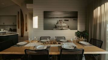 A dining area with a wooden table set for six, surrounded by black chairs. A large painting of a staircase leading to a body of water is on the wall behind the table.