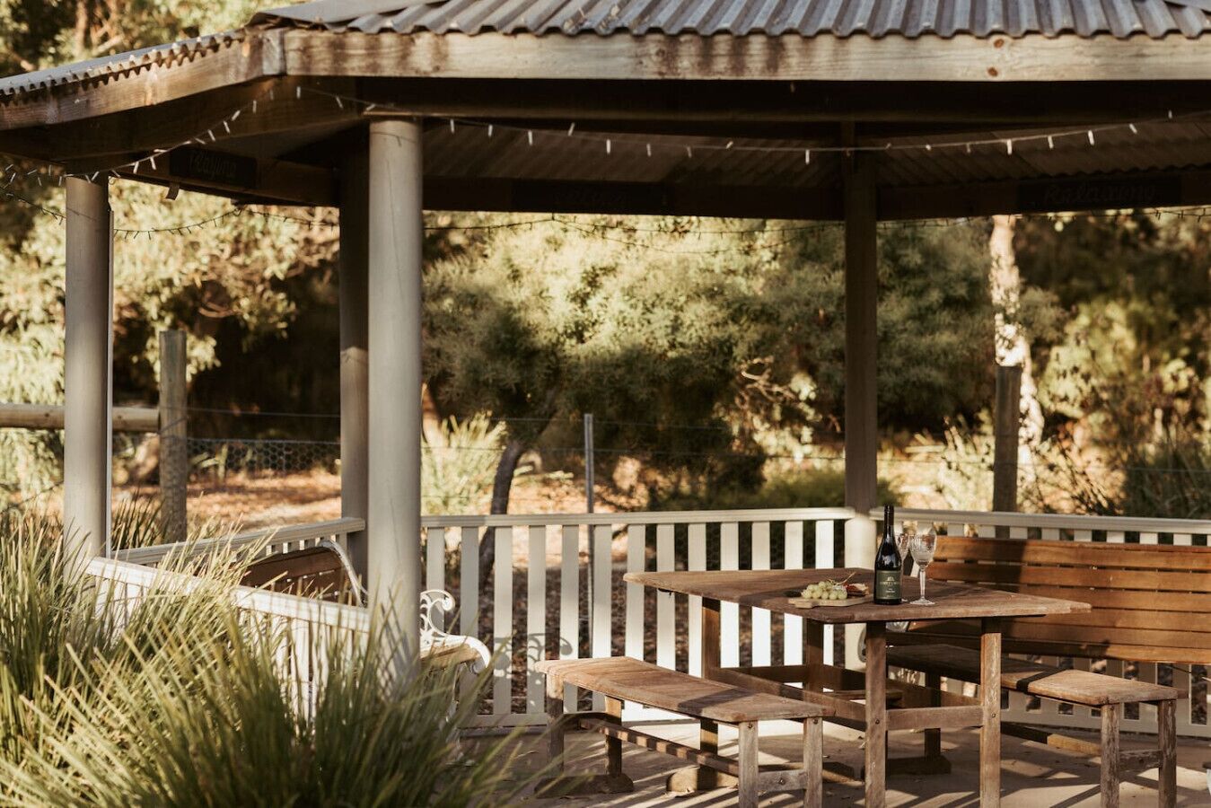 An outdoor patio with wooden benches and a table, surrounded by greenery.
