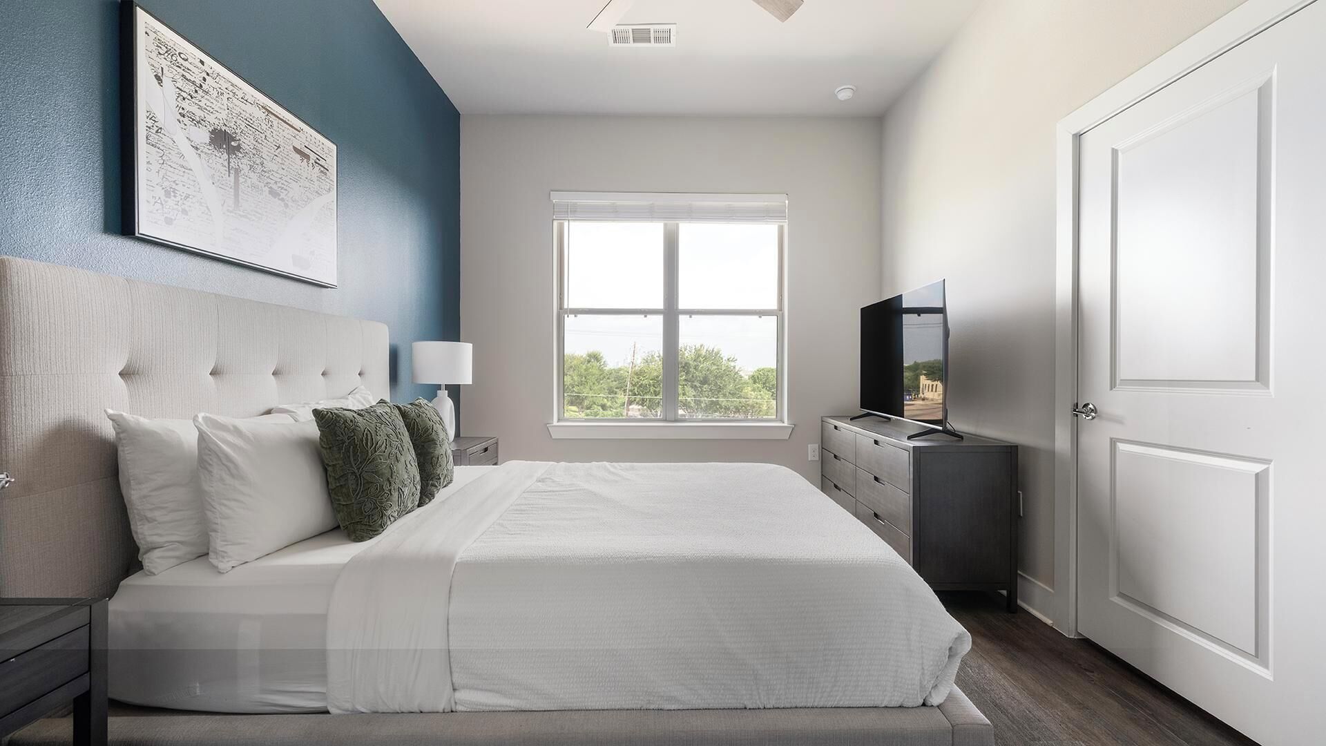 A bedroom with a large bed, a nightstand, a dresser, a TV, and a window.