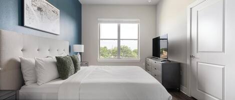 A bedroom with a large bed, a nightstand, a dresser, a TV, and a window.