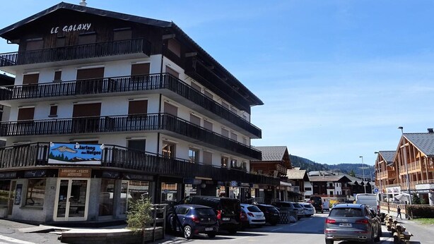 Un hôtel à plusieurs étages doté de balcons, une enseigne indiquant la présence d’une station de ski, et des voitures garées le long de la rue.