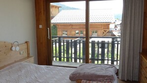 Une chambre à coucher avec un lit, une tête de lit en bois, une table de chevet et une vue sur un bâtiment en bois, visible à travers une porte-fenêtre coulissante.