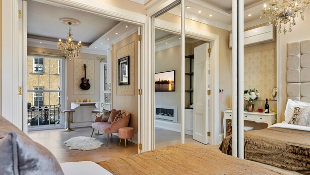 A spacious bedroom with a large bed, a sitting area with a pink sofa, a dining area with a guitar, and a view of the city through a large window. - Apartment in Marylebone with Jacuzzi Bath (London)