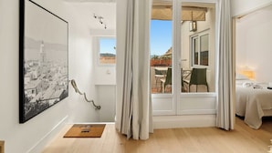 A room with a large framed black and white cityscape painting, a wooden floor, a bed with a white blanket, and a view through a glass door to an outdoor area with a table and chairs. - Magnificent apartment - AC- 2BR- Saint-Tropez (Saint-Tropez)