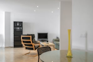 A modern living room with a glass table, a yellow vase, a television, and a bookshelf.