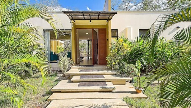 Una casa moderna con un portico coperto, un vialetto che conduce all'ingresso e vegetazione circostante.