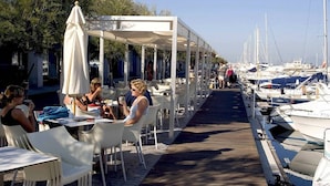 A covered outdoor dining area with white chairs and tables, overlooking a marina with boats.