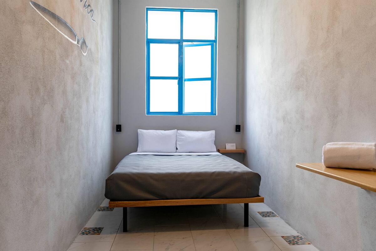 Una habitación estrecha con una cama, una ventana, una toalla en un estante e ilustración de un cuchillo en la pared.