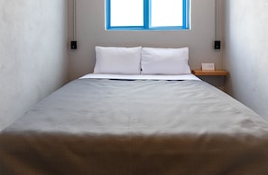 Una cama con ropa blanca, una mesita de noche de madera y un ventanal con marco azul.