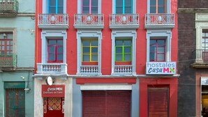 Een gebouw met kleurrijke ramen en balkons, een dichtgeslagen rood luik, en borden met de tekst 'hostales casaMX' en 'belles artes'.