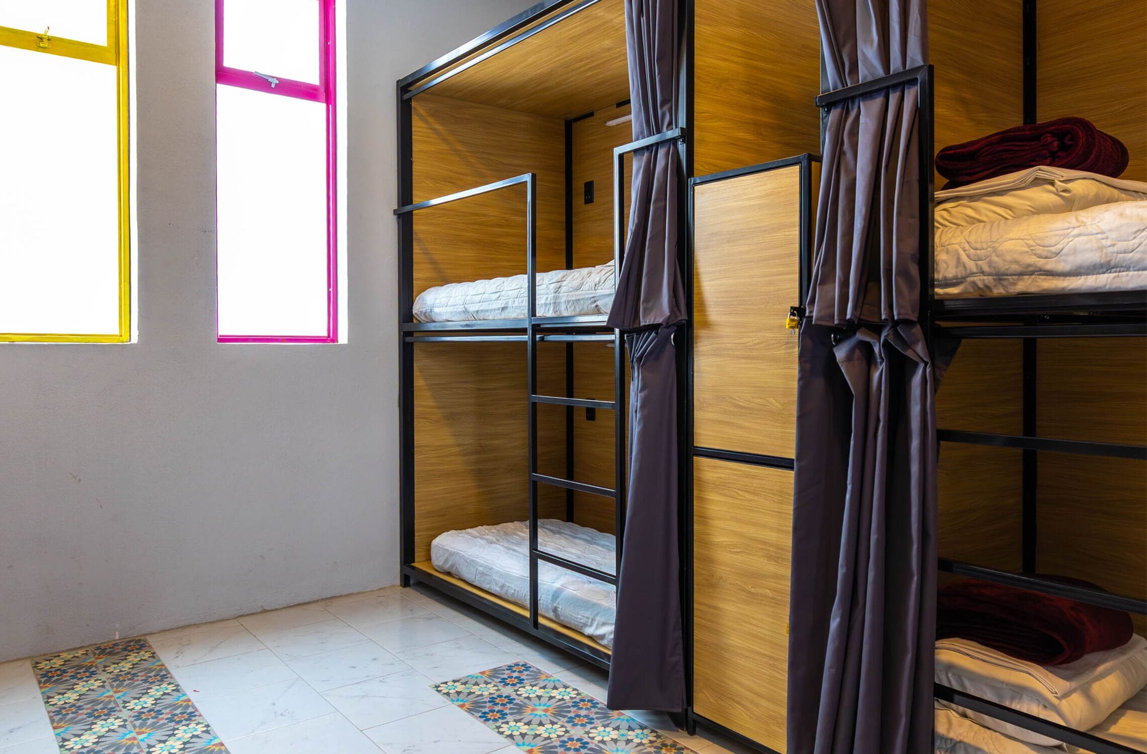 A dormitory room with bunk beds, a patterned rug, and a window with pink framing.
