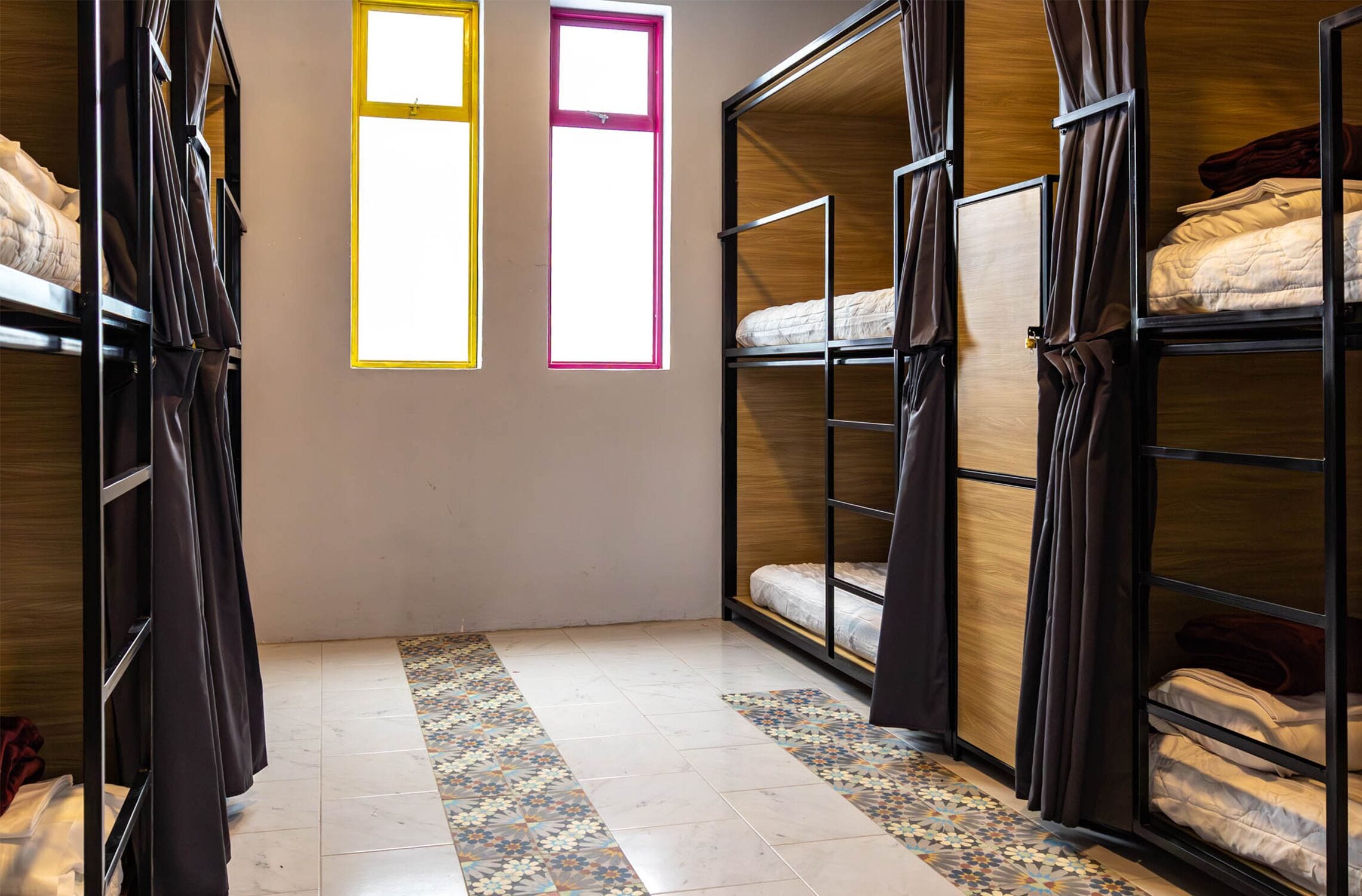 Un dormitorio con literas, suelo estampado y dos ventanas con marcos amarillos y rosados.