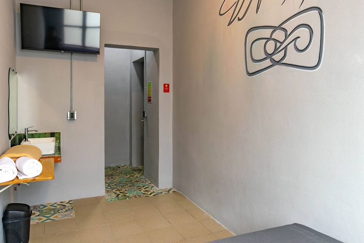 Una habitación de hotel con un televisor montado en la pared, un baño con lavamanos y espejo, un piso con diseño y una puerta que da a otra habitación.