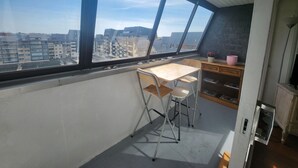 Ein kleiner Balkon mit klappbarem Tisch und Stühlen, einem Holztisch und Blick auf die Stadt durch große Fenster.