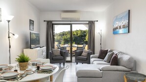 A modern living room with a grey sofa, dining table set with plates and glasses, a TV, and a balcony with a view of greenery. - Casa Colina Sol 611 - Seaview apartment (Carvoeiro)