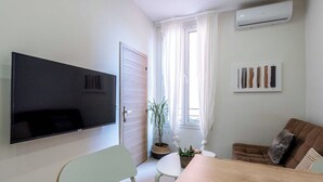 Una sala de estar con un televisor de pantalla plana, una mesa de comedor, sillas, un sofá, una planta en maceta y una ventana con cortinas.