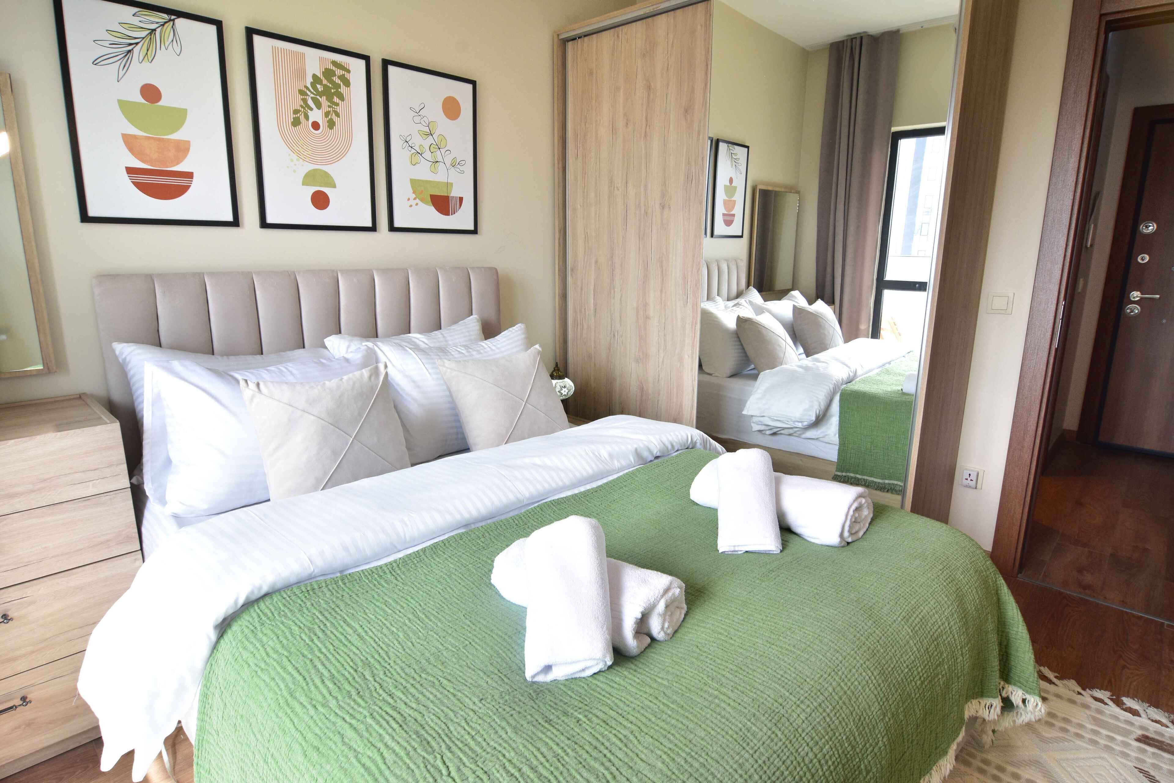 A neatly made bed with white linens, a green throw blanket, and rolled towels. There are three framed artworks on the wall above the bed.