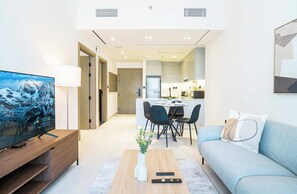 Un salon moderne avec un téléviseur à écran plat, une table basse ornée d’un vase, un canapé et un coin repas avec des chaises, et une cuisine en arrière-plan.