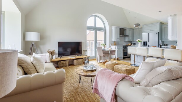 A spacious living room with a television, sofa, and coffee table. A kitchen with white cabinets and a dining area with chairs is visible through an arched window. - Cobblestone Cottage - sleeps 4 guests  in 2 bedrooms (Docking)
