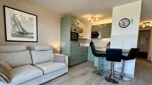 A modern living room with a sofa, a dining area with bar stools, a kitchen with a microwave, and a framed picture on the wall. - 🏖️ Les Pieds dans le Sable - Unique Apartment with Direct Beach Access (Le Crotoy)