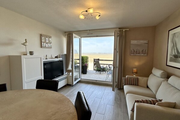 A hotel room with a TV, a sofa, a dining table, and a balcony with a view of the beach.