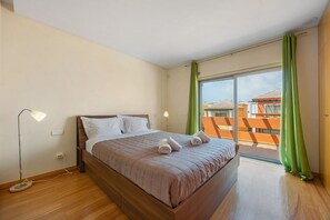 Une chambre à coucher avec un lit en bois, une table de chevet, une lampe et une vue sur les immeubles par une grande fenêtre.