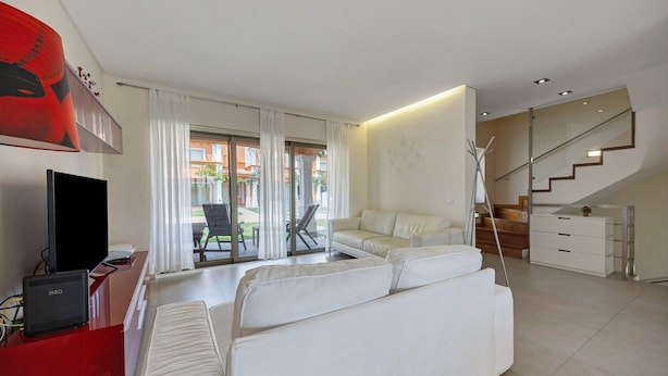 A modern living room with a sofa, a TV, a staircase, and a view of the outdoors. - Holiday Home 'Orange Lux Tavira Residence 3I' with Shared Pool, Wi-Fi and Air Conditioning (Luz de Tavira)
