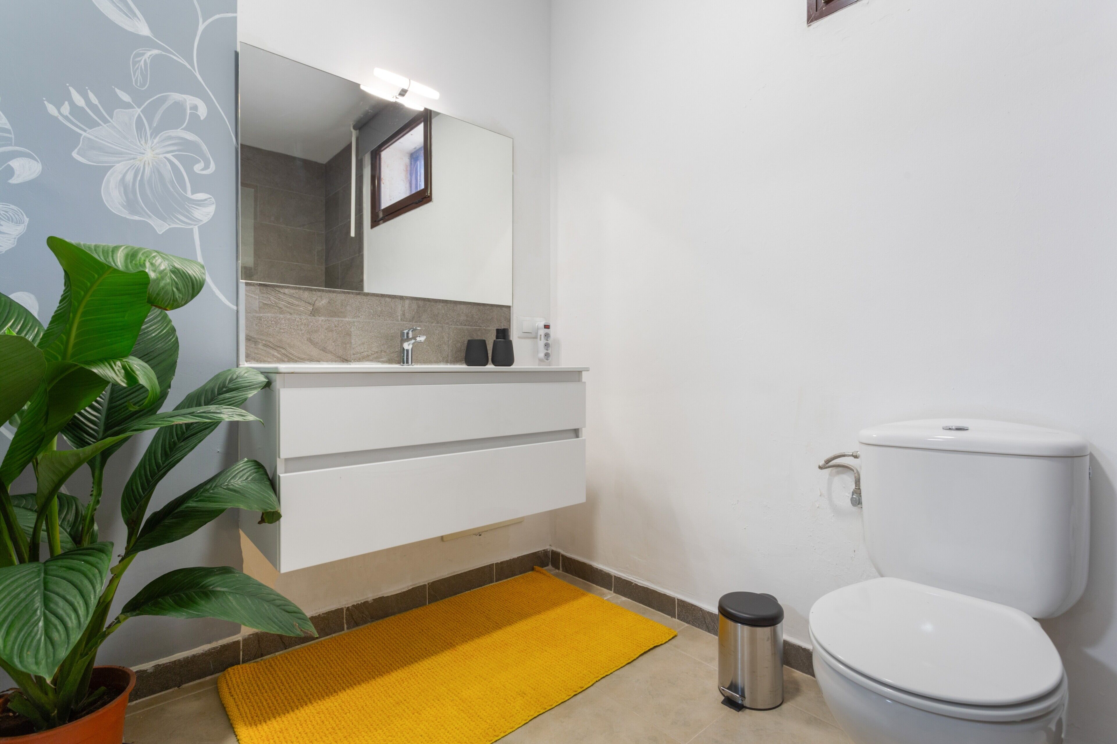 Une salle de bain avec un lavabo blanc, des toilettes, un tapis jaune et une plante en pot.