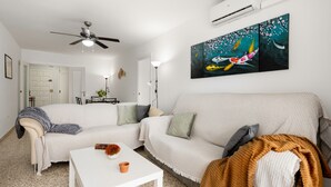Una sala de estar con dos sofás blancos, una mesa de centro blanca, un ventilador de techo y un cuadro en la pared.