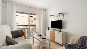 Una sala de estar con un sofá, una mesa de centro, un televisor sobre un mueble de madera y un balcón con vistas a edificios.