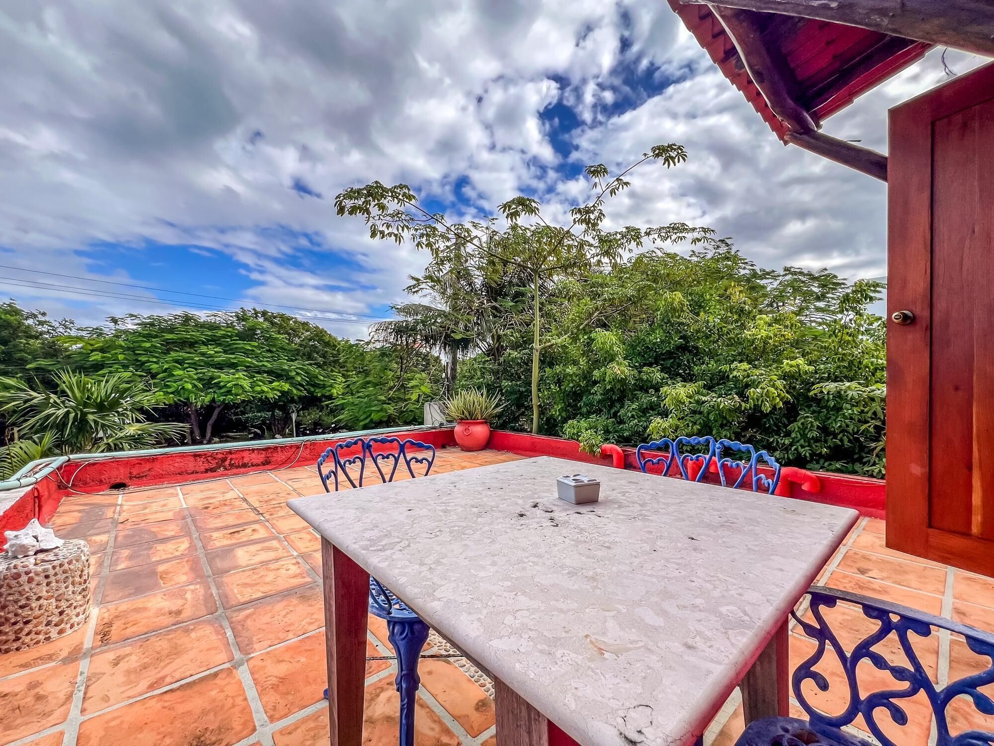 Une terrasse avec une table et des chaises, entourée d’un mur rouge et offrant une vue sur une végétation luxuriante sous un ciel partiellement nuageux.
