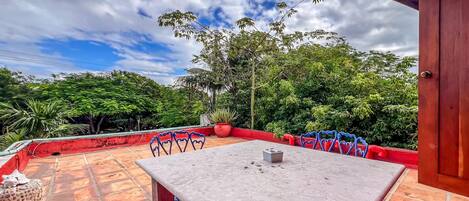 Une terrasse avec une table et des chaises, entourée d’un mur rouge et offrant une vue sur une végétation luxuriante sous un ciel partiellement nuageux.