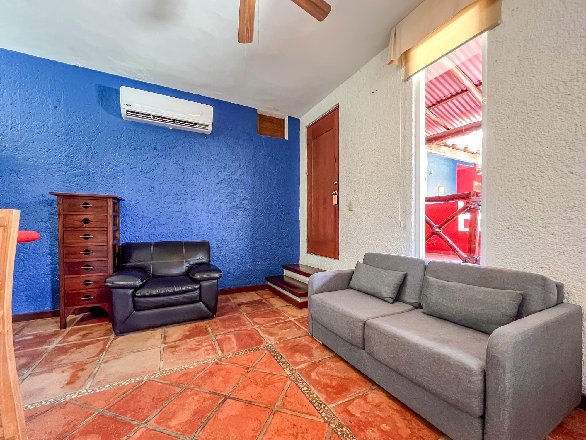 Un salon avec un mur bleu, un fauteuil en cuir noir, un canapé gris, un meuble en bois et un sol carrelé.