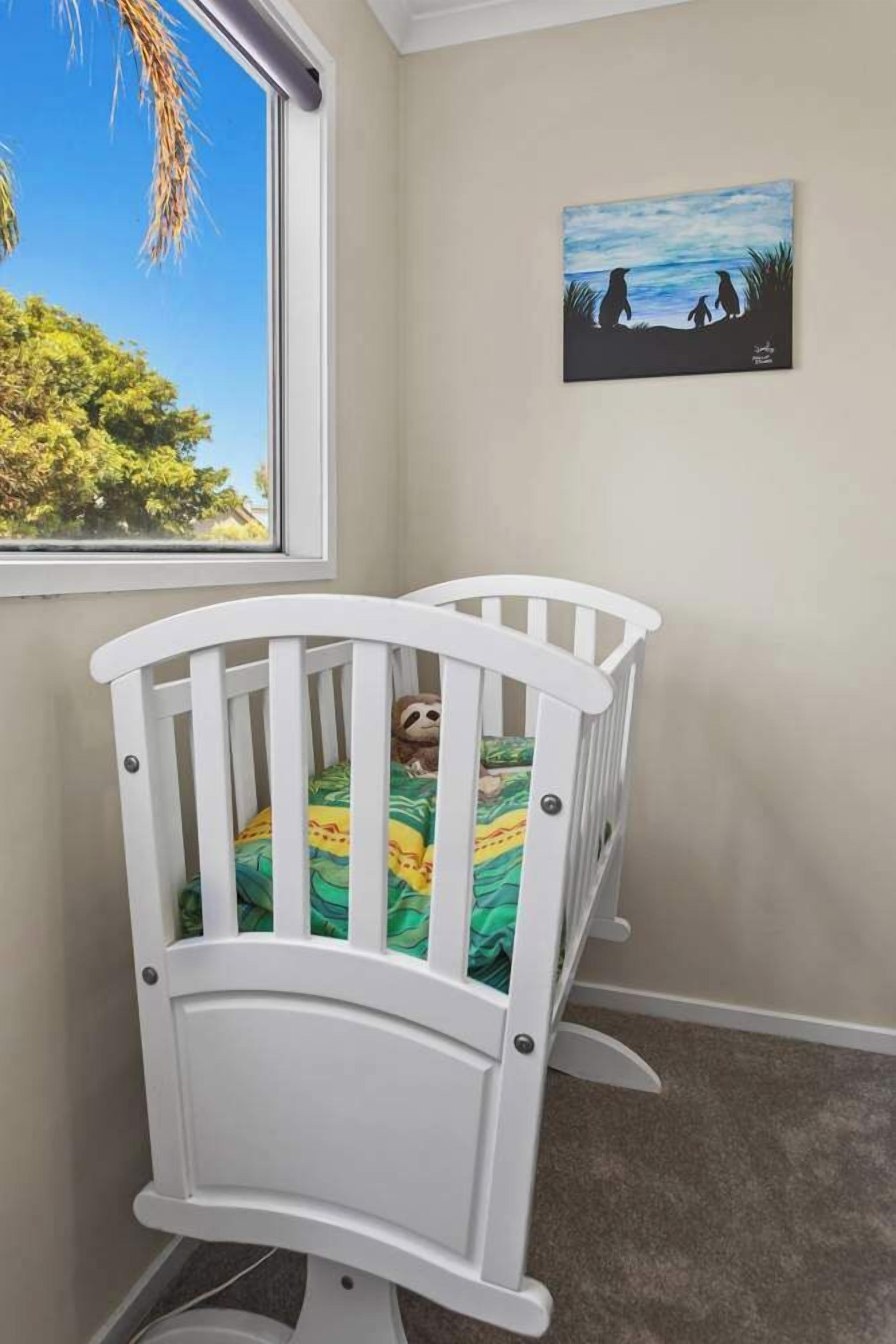 A white crib with a green and yellow patterned blanket, a painting of penguins on the wall, and a window with a view of trees and sky.