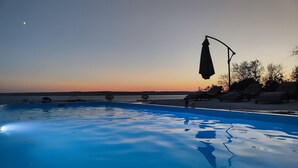 A swimming pool with lounge chairs and umbrellas, surrounded by trees and a sunset view.