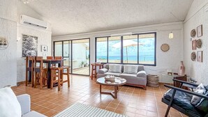 A spacious living area with a sofa, coffee table, and dining set. Large windows offer a view of the beach. - Ocean View PH in Punta Sur (Isla Mujeres)