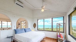 A bedroom with a bed, a ceiling fan, a window with a view of the ocean, and a small table. - Ocean View PH in Punta Sur (Isla Mujeres)