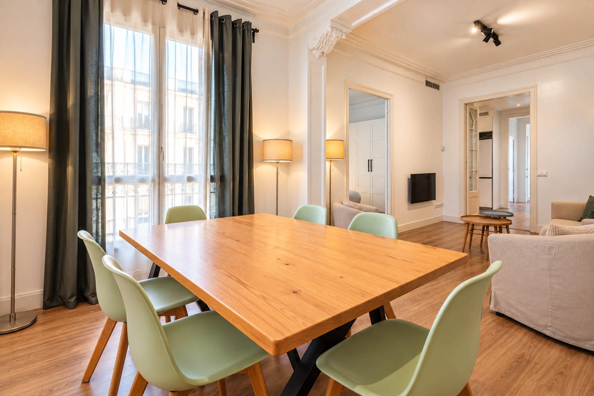 A dining area with a wooden table and green chairs, a sofa, and a TV in a modern apartment.