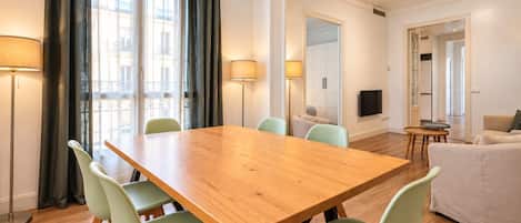 A dining area with a wooden table and green chairs, a sofa, and a TV in a modern apartment.