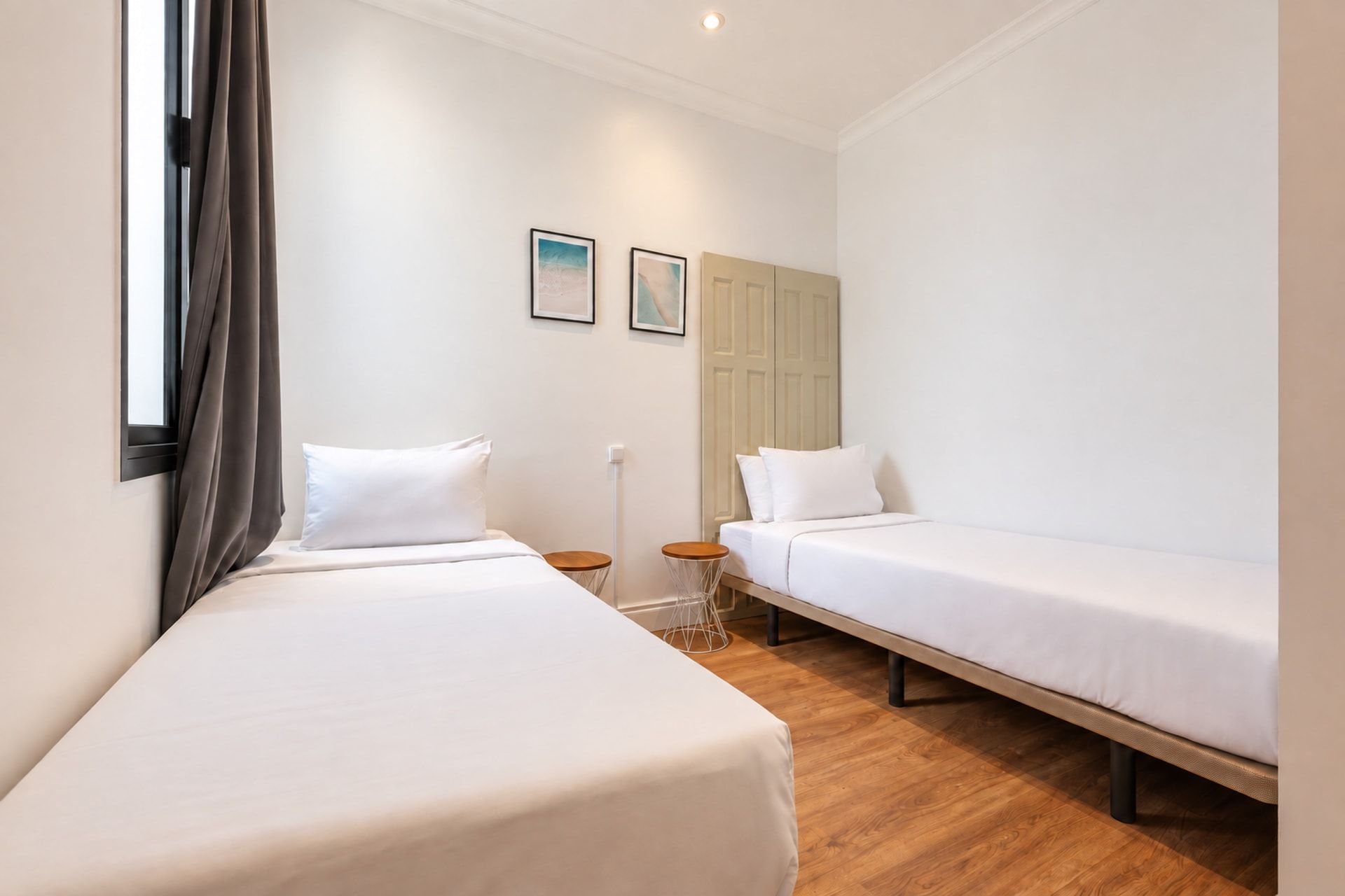 A hotel room with two single beds, a wooden floor, a window with curtains, and a door.