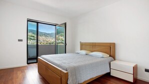Un dormitori amb un llit de fusta, una taula de nit i una porta corredissa de vidre que dona a un balcó amb vista a la ciutat.