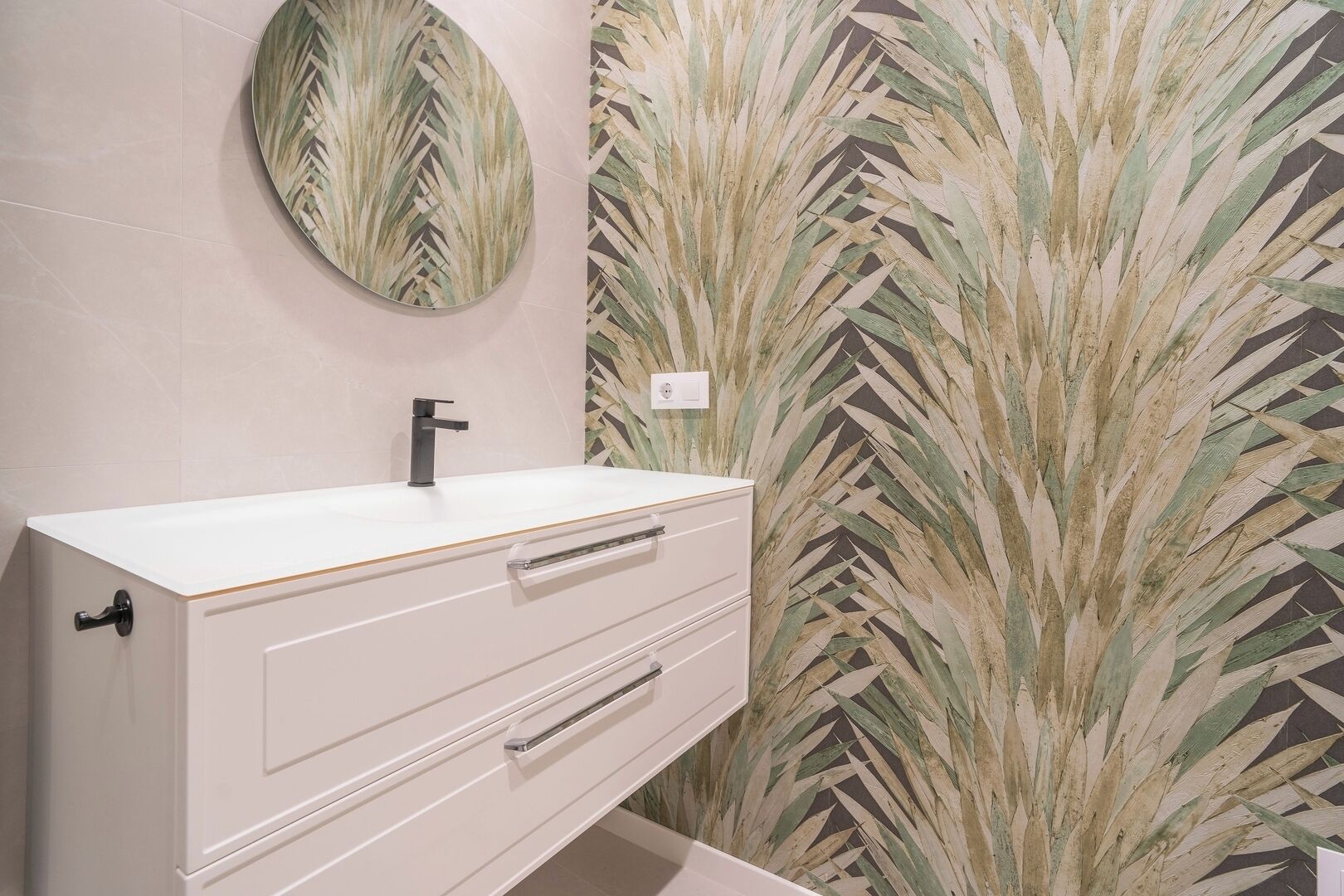 A bathroom with a white vanity, a black faucet, and a round mirror. The wall features a large, leafy pattern.