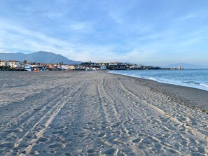 Une plage où l’on voit des empreintes de pas menant à l’eau, un ciel dégagé et des bâtiments en arrière-plan.
