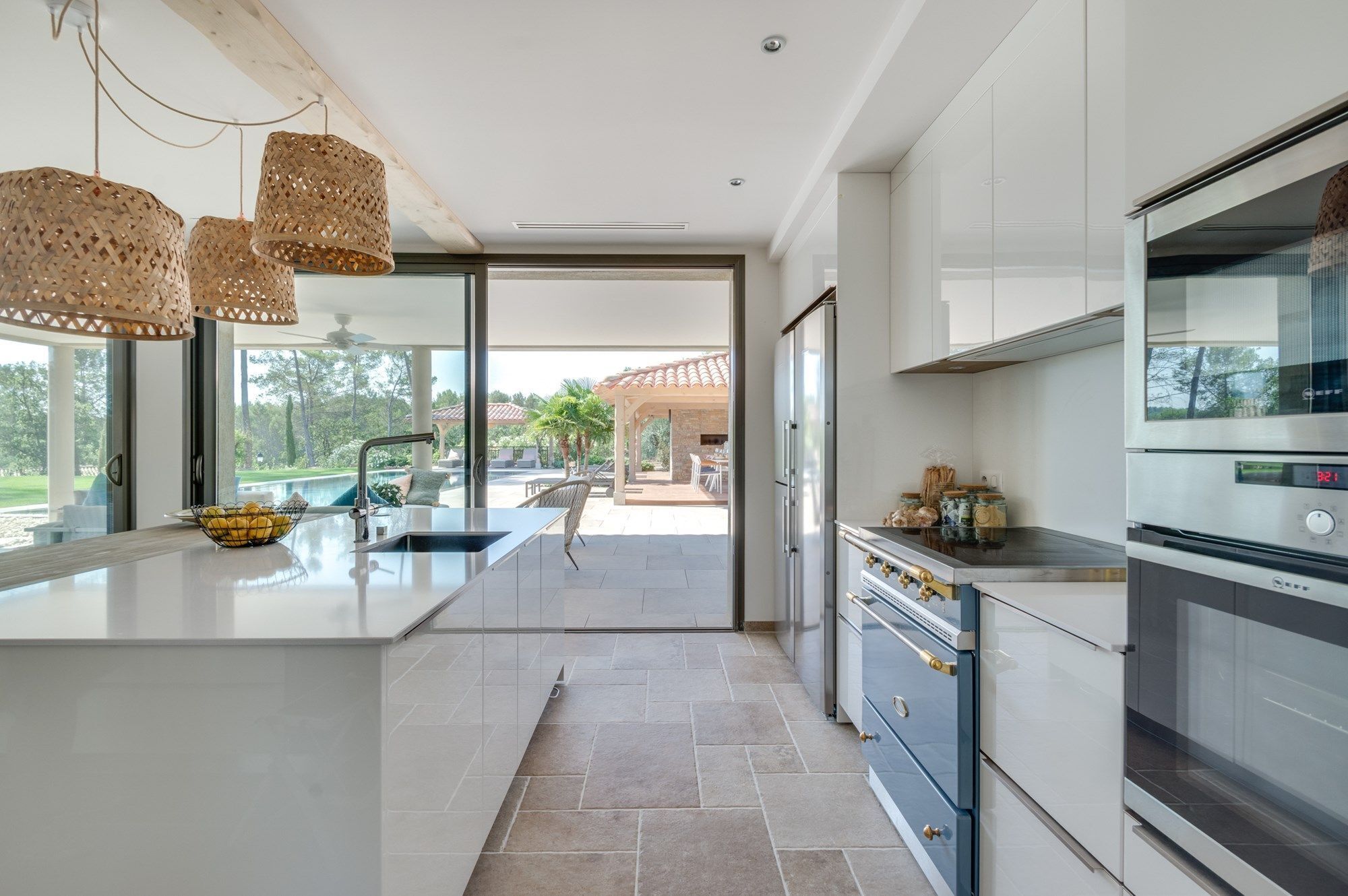 Une cuisine moderne dotée d’un grand îlot central, d’appareils électroménagers en acier inoxydable et offrant une vue sur un patio avec coin salon extérieur.