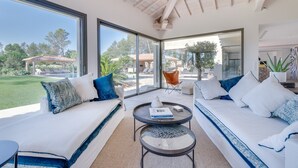 Een moderne woonkamer met een glazen wand, twee witte banken met blauwe accenten, een ronde salontafel en uitzicht op een zwembad en tuin.