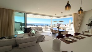 A modern living room with a sofa, dining table, and chairs, large windows with a view, and pendant lighting.